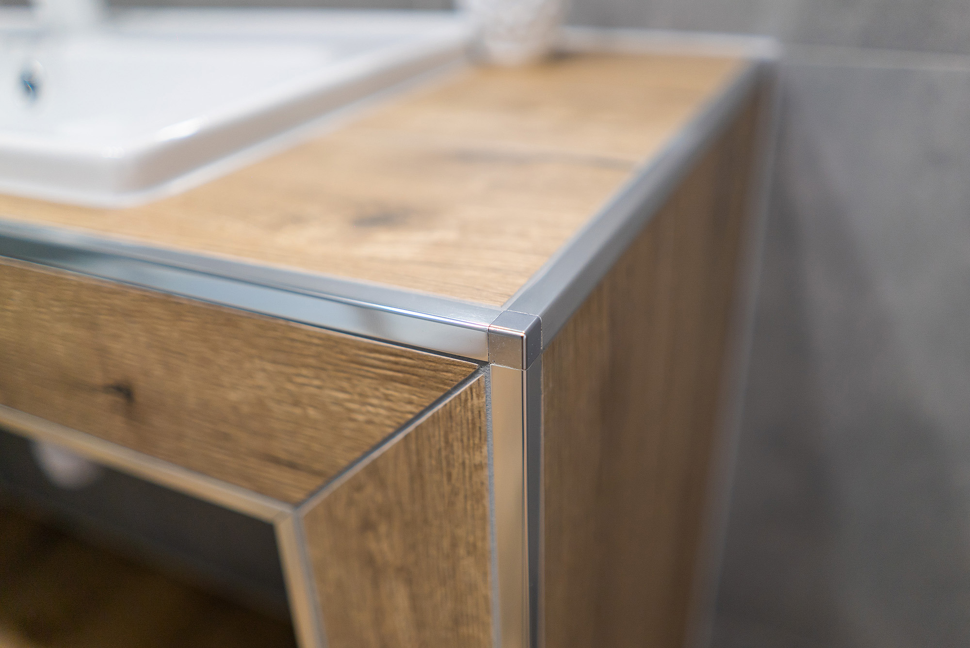 Blat łazienkowy z drewnopodobnym dekorem i srebrnymi listwami wykończeniowymi, narożnik blatu z umywalką w tle.