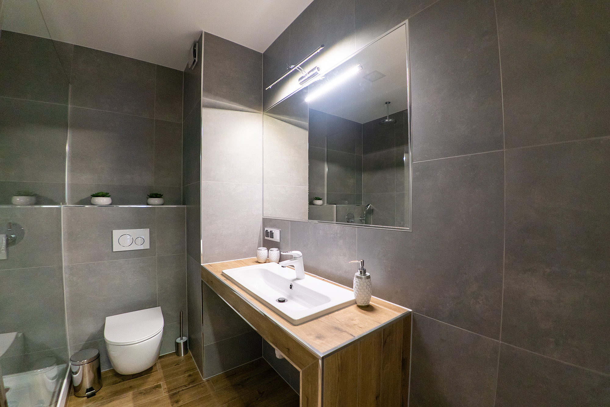 Nowoczesna łazienka z szarymi płytkami, podwieszaną toaletą, drewnianym blatem pod umywalką i dużym lustrem z oświetleniem LED. Na półce nad toaletą widoczne małe rośliny w białych doniczkach.