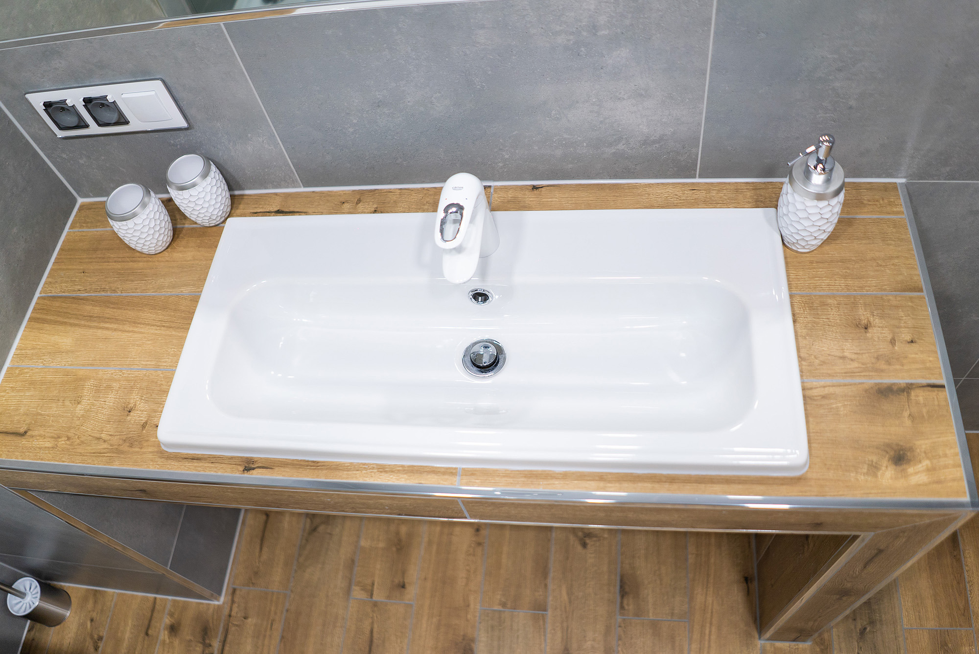 Nowoczesna umywalka w łazience z blatem imitującym drewno, biała armatura Grohe, szare płytki i dekoracyjne akcesoria łazienkowe.