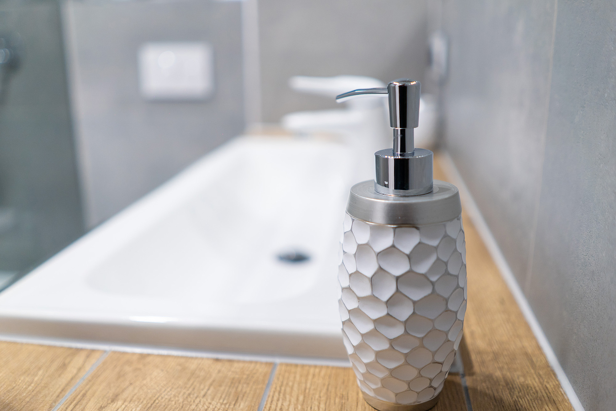 Biały dozownik do mydła z geometrycznym wzorem plastra miodu, umieszczony na drewnianym blacie w łazience z umywalką w tle.