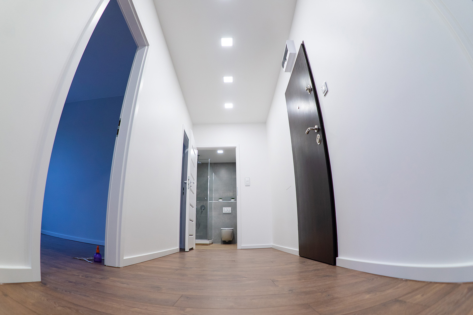 Korytarz z trzema otworami drzwiowymi, widok na wyremontowaną łazienkę z kabiną prysznicową i podwieszaną toaletą, brązowe panele podłogowe, białe ściany i sufit z wbudowanymi lampami.