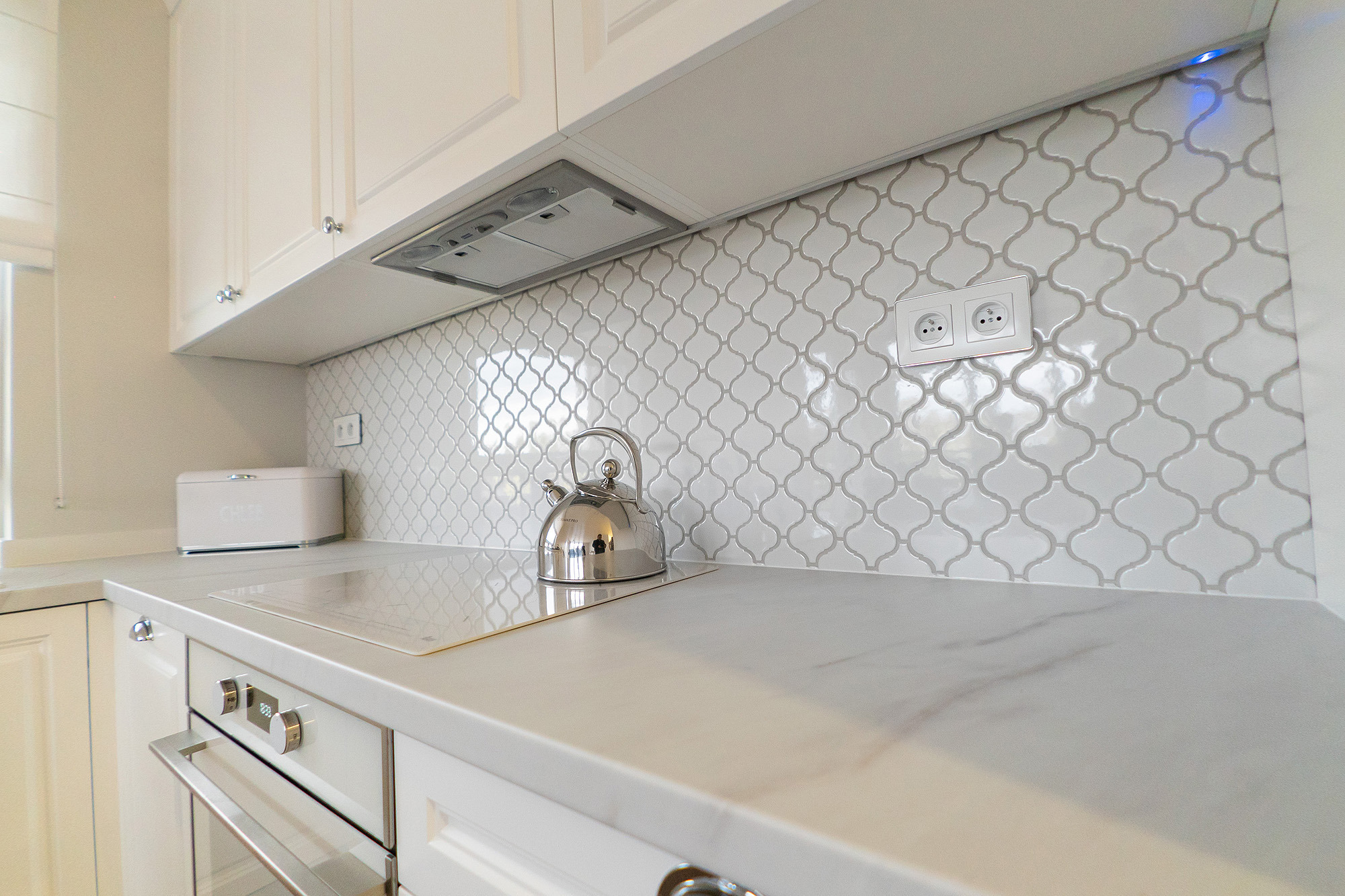 Biała kuchnia z błyszczącymi płytkami arabeska na ścianie, chromowany czajnik i piekarnik w zabudowie.
