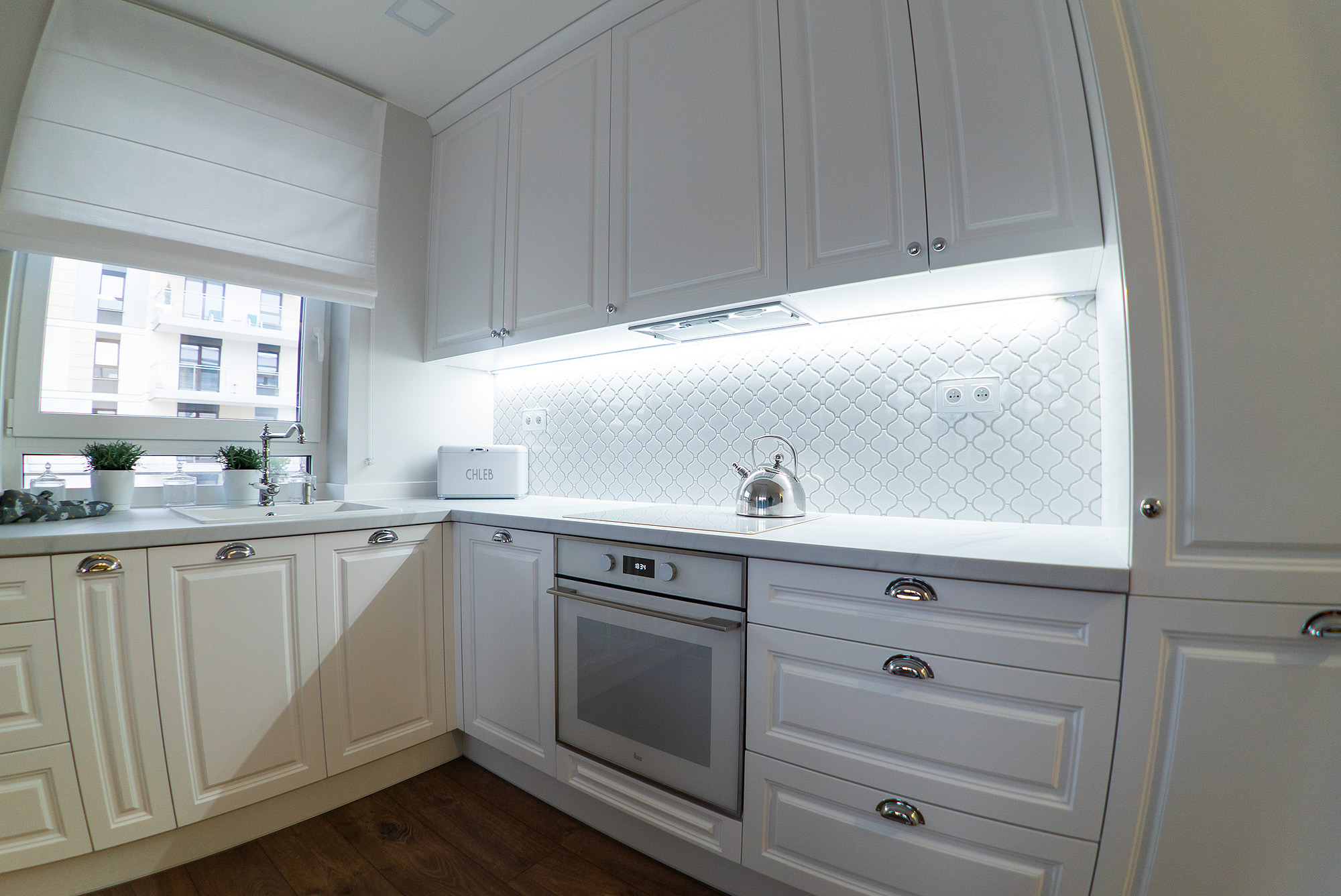 Jasna kuchnia z białymi szafkami, mozaiką nad blatem, srebrnym czajnikiem i piekarnikiem marki Teka.