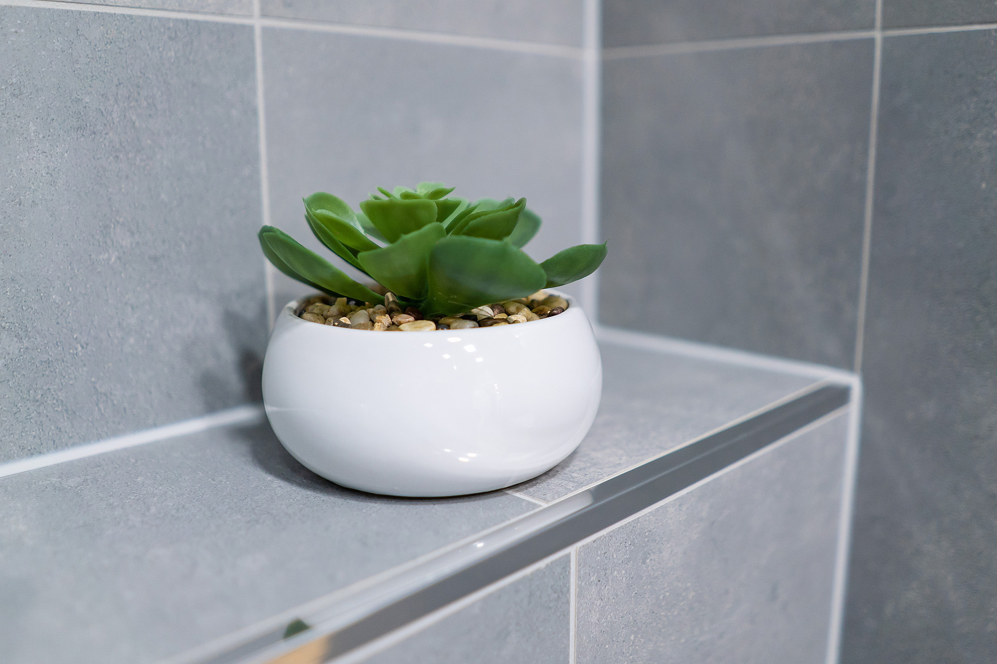 Sztuczny sukulent w białej, okrągłej doniczce na półce wyłożonej szarymi płytkami w łazience.