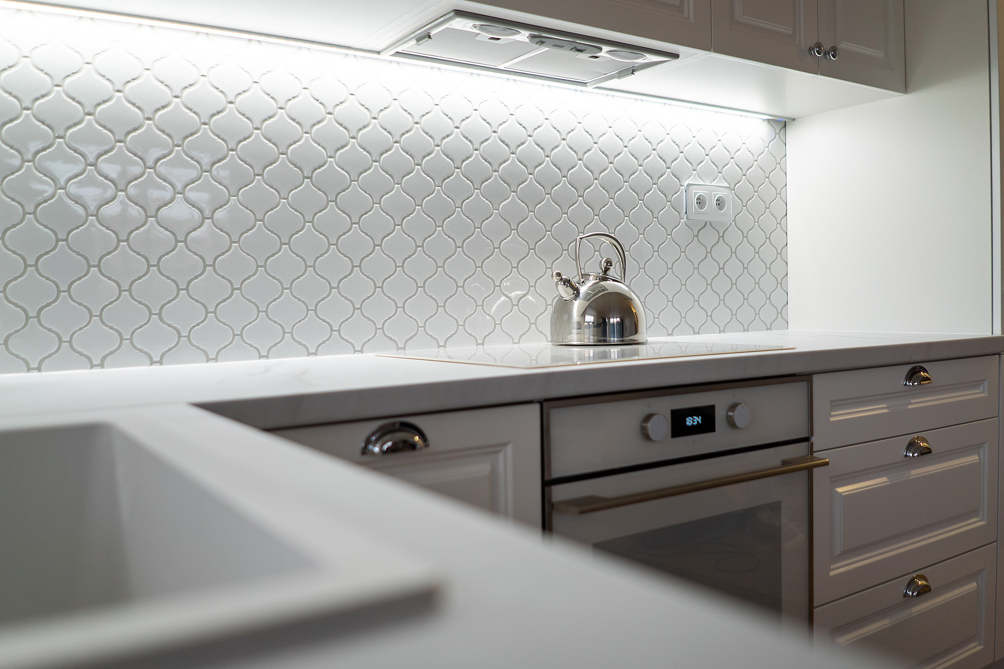 Biała kuchnia z błyszczącymi płytkami w marokański wzór, czajnik na płycie indukcyjnej i zabudowany piekarnik z wyświetlaczem.