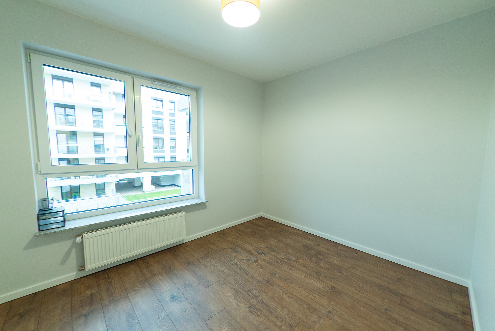 Pusty pokój z panelami podłogowymi w odcieniach brązu, białymi ścianami, grzejnikiem pod oknem i widokiem na budynek mieszkalny z balkonami.