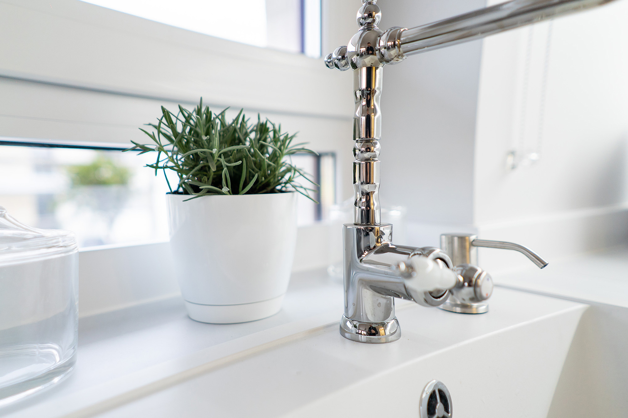 Bateria umywalkowa w stylu retro z białymi kurkami, obok doniczka z ziołami na tle okna w jasnej łazience.