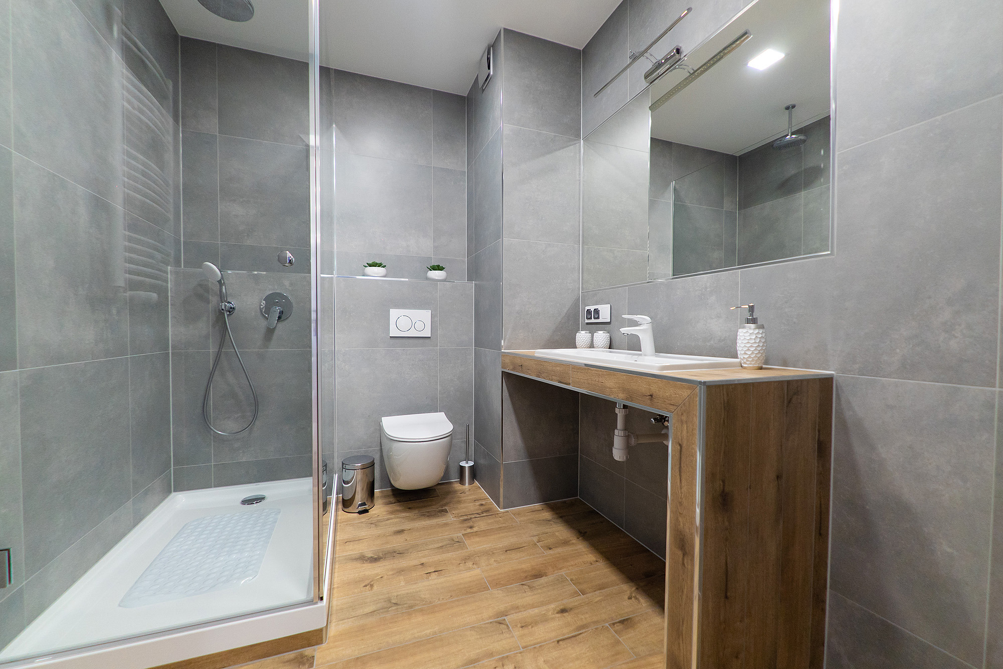 Nowoczesna łazienka z szarymi płytkami, drewnopodobną podłogą i kabiną prysznicową typu walk-in, minimalistyczna umywalka z blatem z efektem drewna, toaleta podwieszana z minimalistycznym przyciskiem.