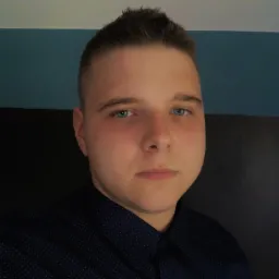 Portret młodego mężczyzny w ciemnej koszuli z drobnym wzorem, z niebieskimi oczami i krótkimi włosami, na tle dwukolorowej ściany.