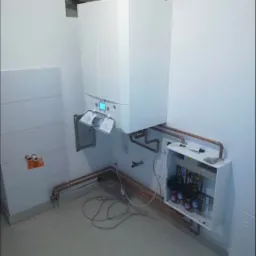 Nowo zainstalowany, biały piec gazowy wiszący na ścianie, z otwartą klapką i widocznym wyświetlaczem, podłączony miedzianymi rurami do instalacji w ścianie i skrzynki z zaworami.