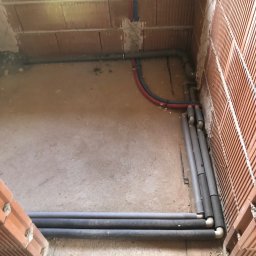 FHU - Surowe wnętrze z ceglanymi ścianami w trakcie budowy, widoczne rury instalacyjne w izolacji, prowadzone wzdłuż ścian i pod podłogą, z elementami instalacji wodnej wystającymi ze ściany.