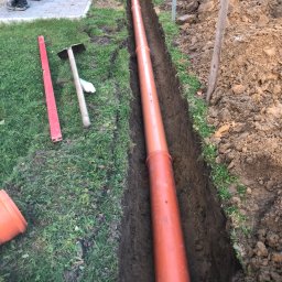 FHU - Pomarańczowa rura kanalizacyjna ułożona w wykopie ziemnym, widoczny poziomica, szpadel i narzędzia na trawie obok wykopu.