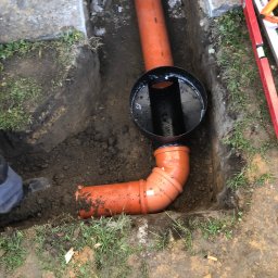 FHU - Pomarańczowe rury kanalizacyjne z czarną studzienką rewizyjną w wykopie, widoczny fragment poziomicy i but pracownika.