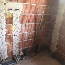 FHU - Surowa ściana z czerwonej cegły z wyżłobieniami wypełnionymi zaprawą, widoczne rury instalacji w szarej izolacji i pionowa rura kanalizacyjna, żółte oznaczenia na ścianie.