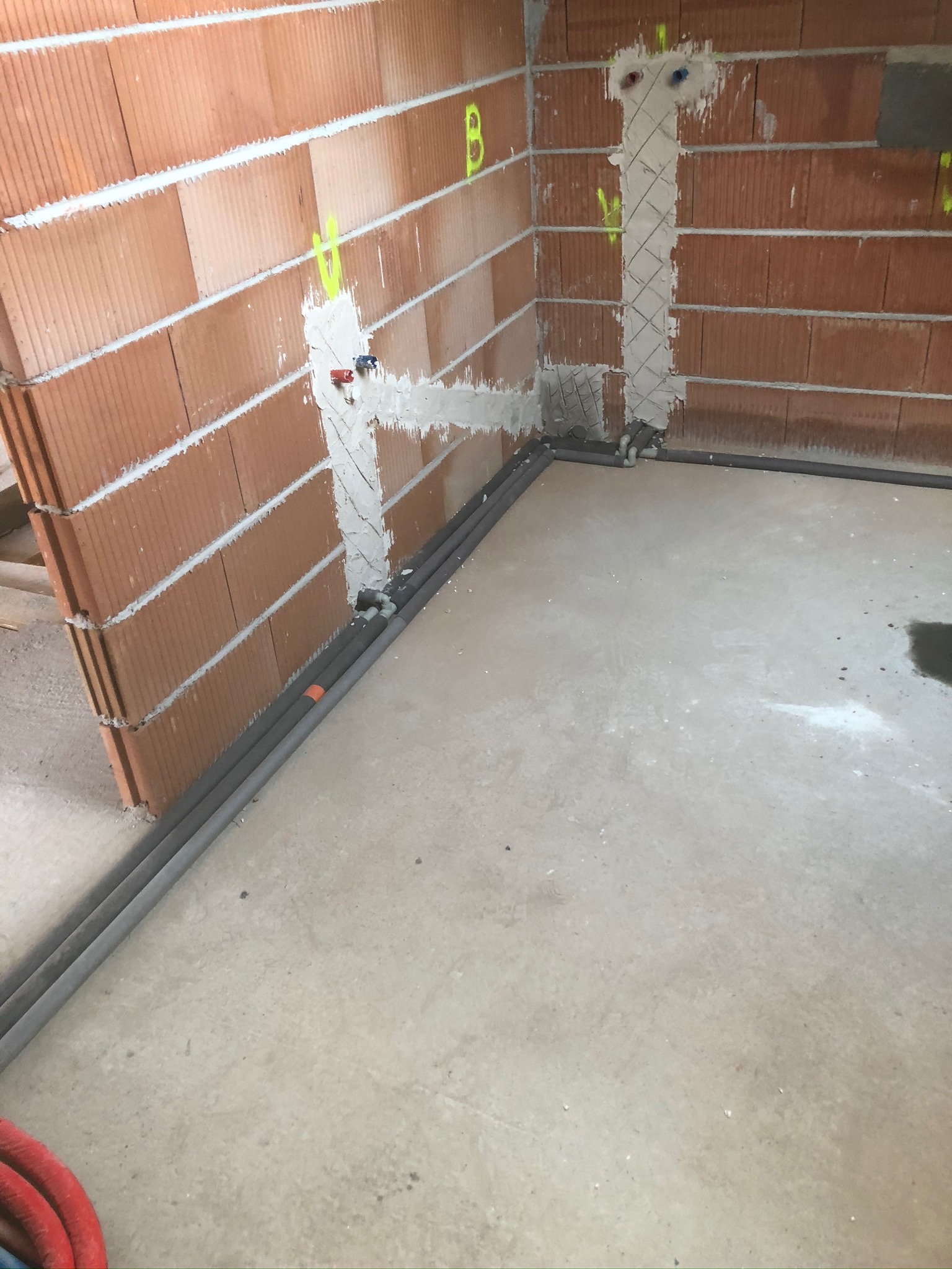 Świeżo położone rury w ścianie z pustaków, zabezpieczone szpachlą, z widocznymi oznaczeniami i fragmentem czerwonego węża na podłodze.