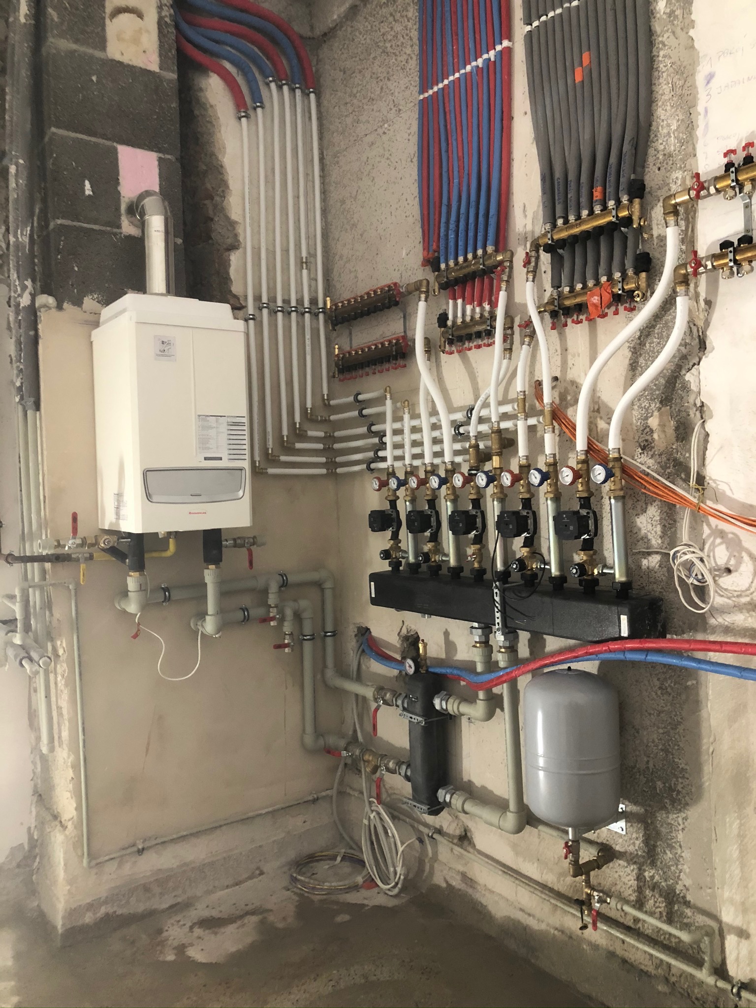 Nowoczesny, biały piec gazowy wiszący na ścianie w surowym stanie, z widocznymi rurami instalacji grzewczej i rozdzielaczami podłogówki, w trakcie podłączania, z widocznym szarym zbiornikiem...