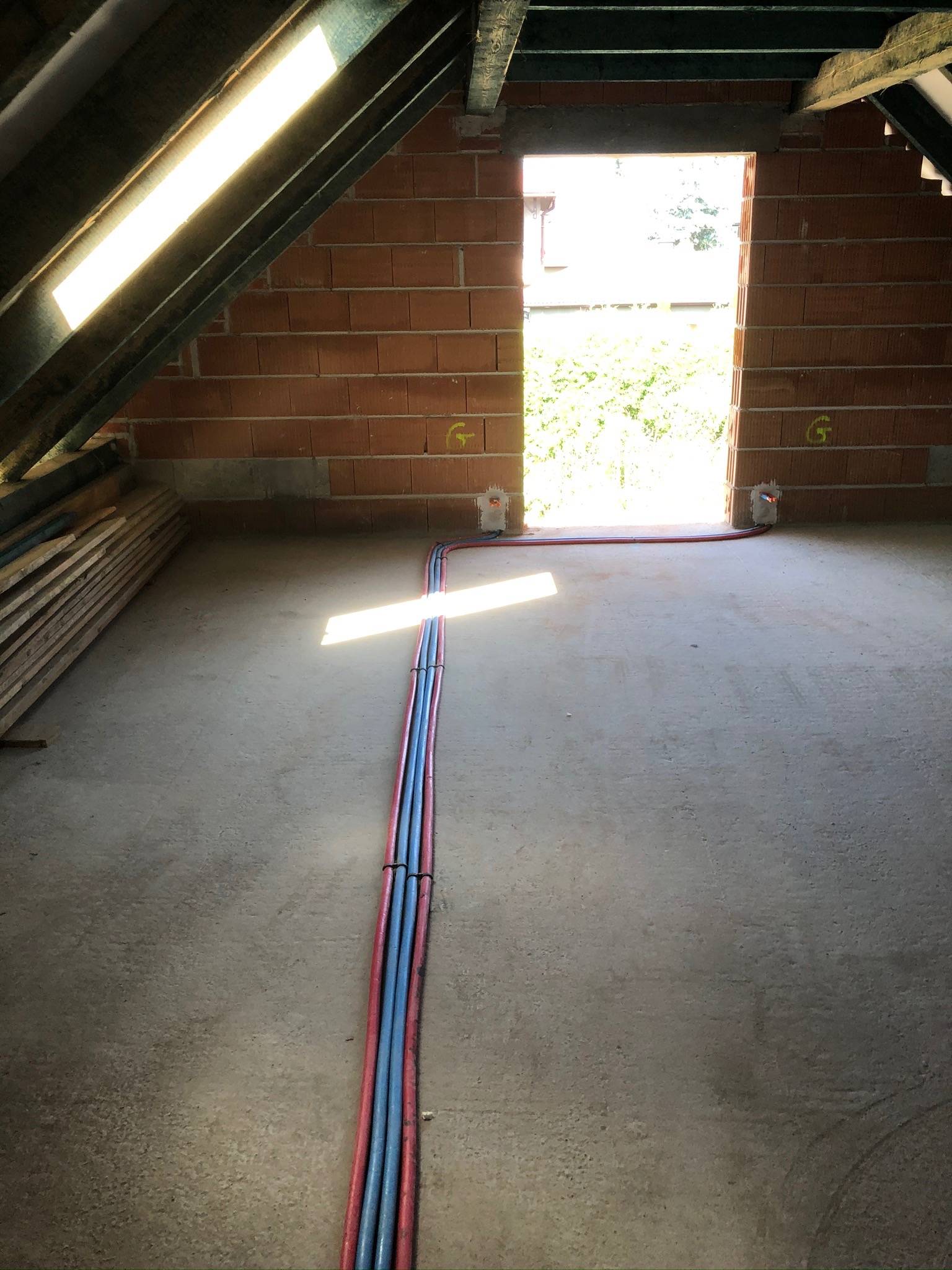 Surowy stan poddasza z ceglanymi ścianami i wiązką rur instalacyjnych w kolorze czerwonym i niebieskim, biegnącą wzdłuż betonowej podłogi w kierunku otworu drzwiowego w głębi kadru, z widocznym...