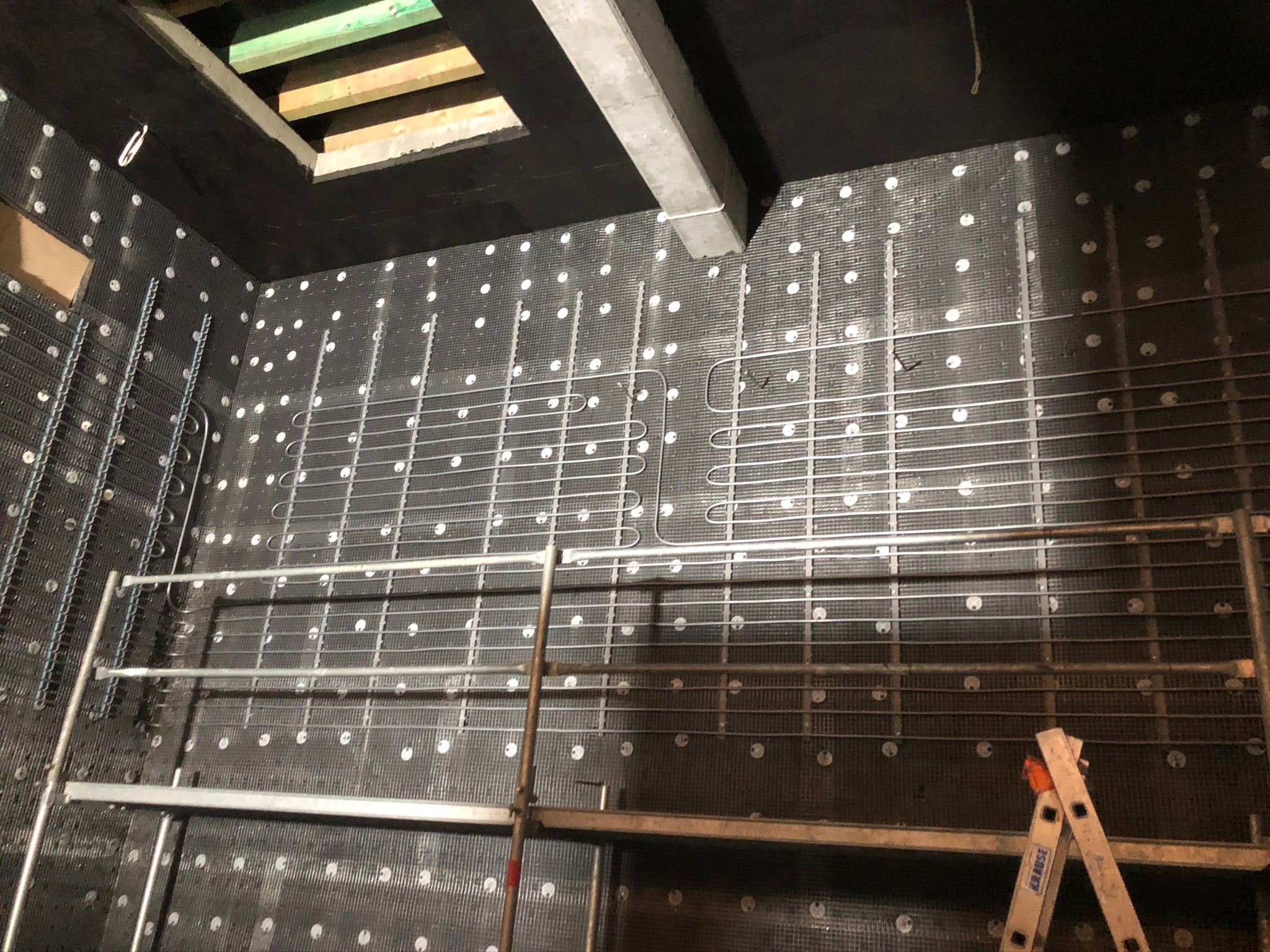 Montaż ogrzewania podłogowego ściennego: rury rozprowadzone na czarnej macie izolacyjnej z białymi punktami, widoczne rusztowanie i drabina Krause.