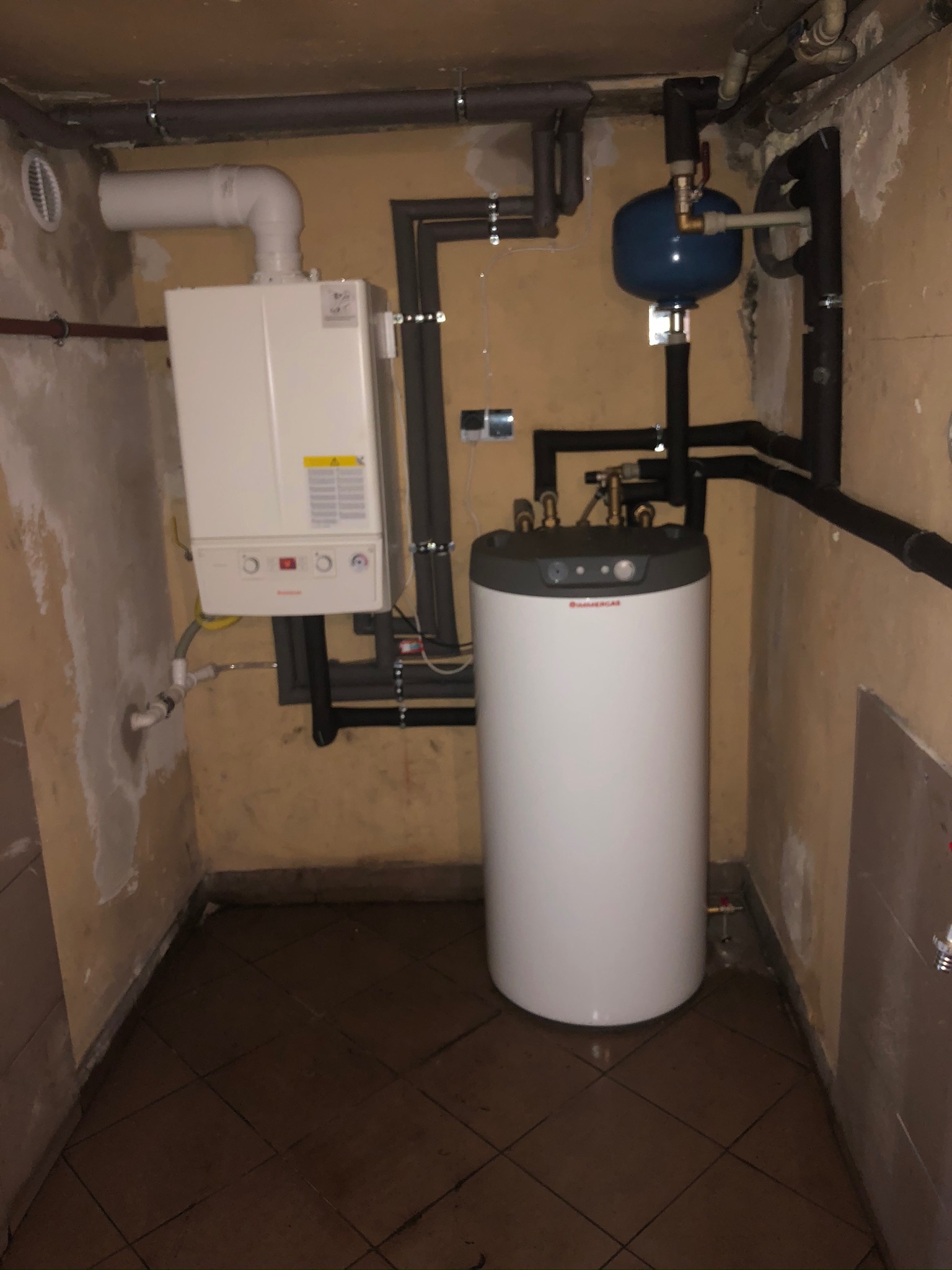 Kompaktowa kotłownia w piwnicy z białym piecem gazowym i zasobnikiem ciepłej wody Dimmergas, połączonymi czarnymi rurami, oraz niebieskim zbiornikiem wyrównawczym, na tle surowych ścian i brązowej...