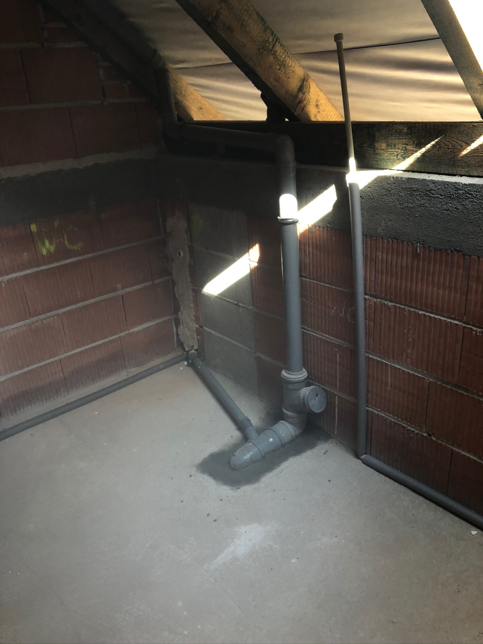 Surowe wnętrze poddasza z widoczną instalacją kanalizacyjną z szarych rur PCV, świeżo wmurowanych w czerwoną cegłę, z fragmentami zaprawy i betonowej podłogi w promieniach słonecznych.