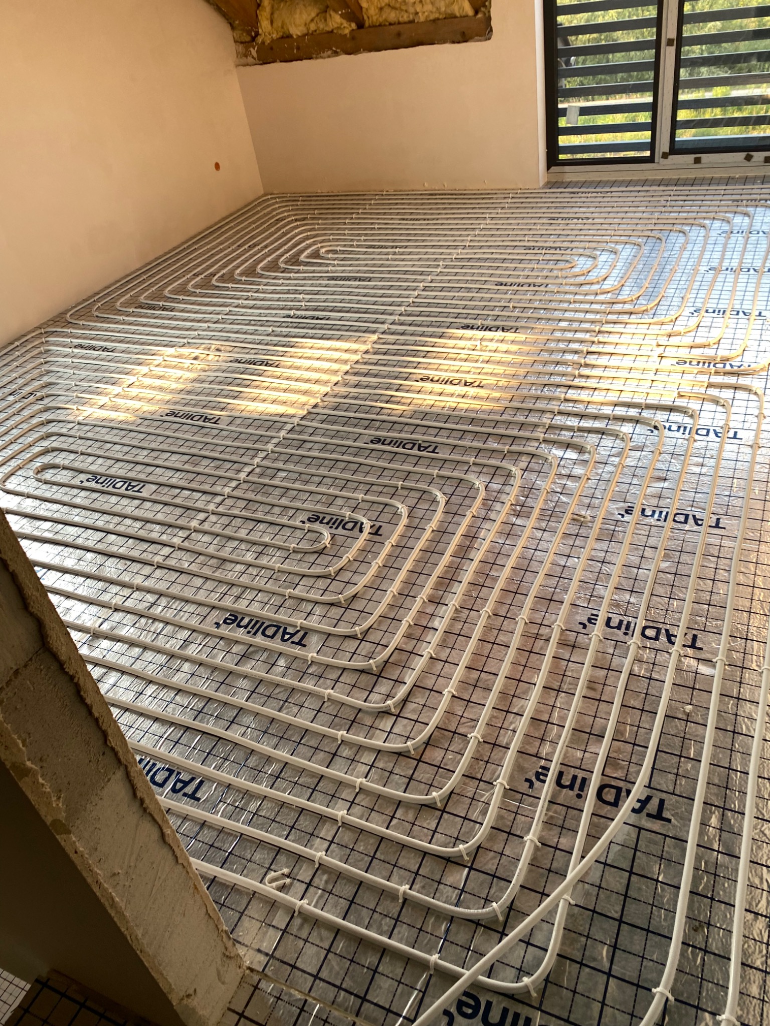 Białe rury ogrzewania podłogowego ułożone spiralnie na folii z nadrukowaną siatką i logotypem TADIINE, widok z góry na poddaszu w trakcie instalacji.
