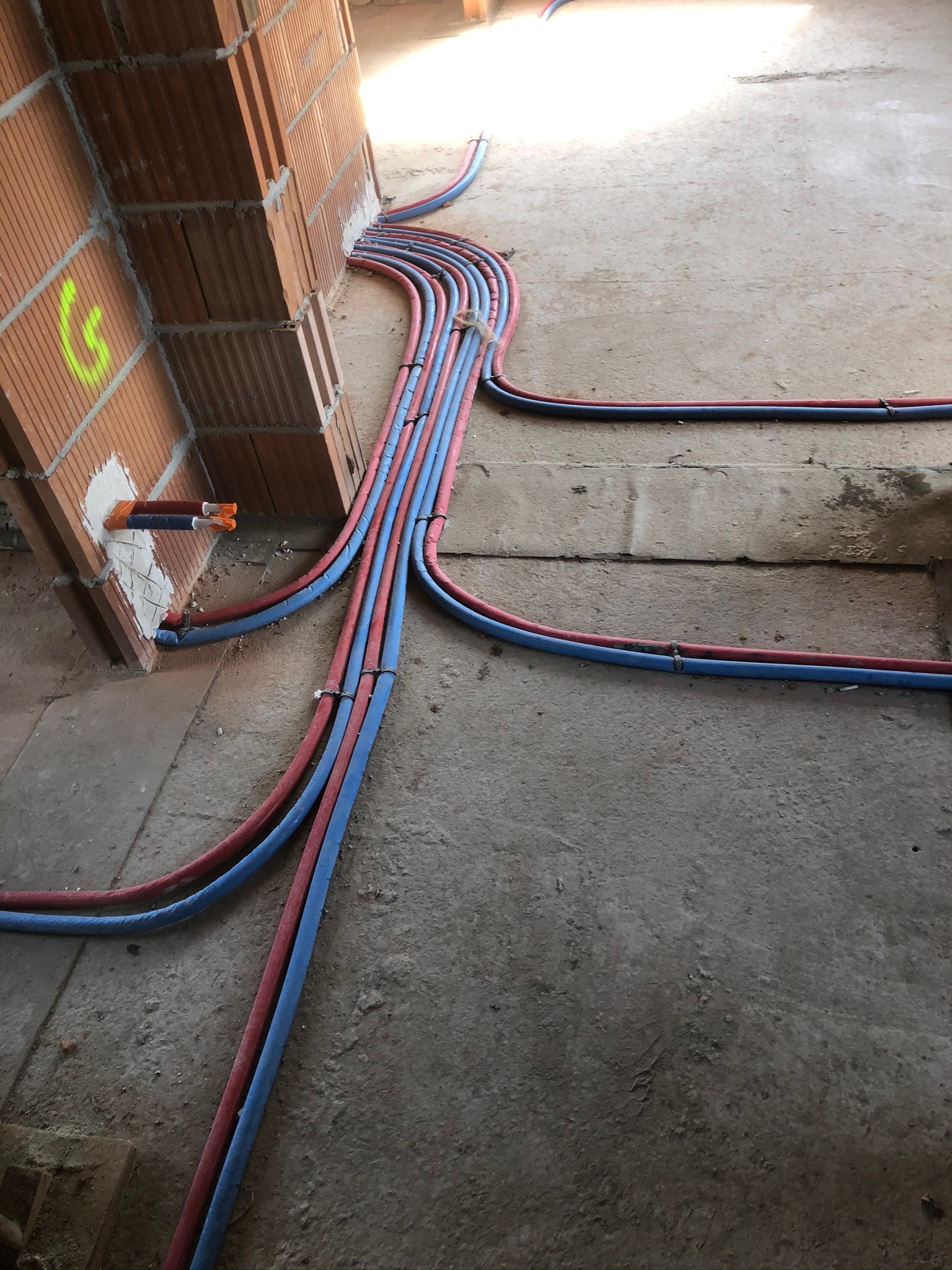 Wczesny etap układania instalacji ogrzewania podłogowego w Krakowie: liczne rury w kolorach czerwonym i niebieskim wychodzą ze ściany z cegły, biegnąc po surowej posadzce.