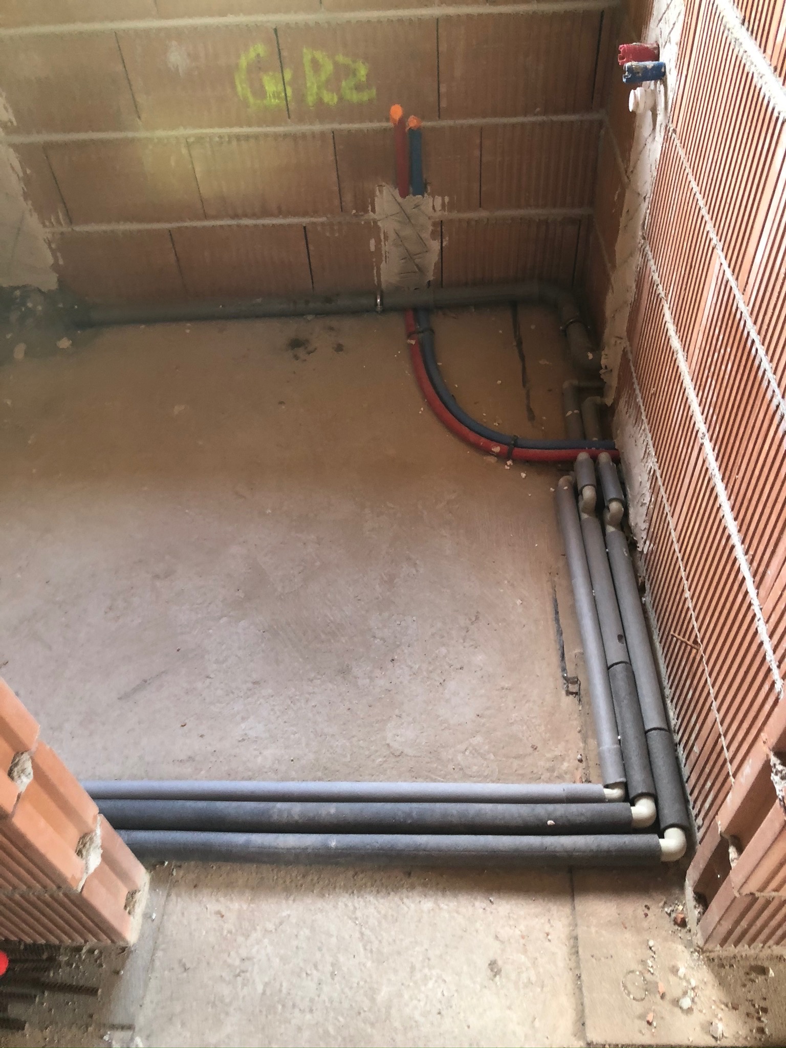Surowe wnętrze z ceglanymi ścianami w trakcie budowy, widoczne rury instalacyjne w izolacji, prowadzone wzdłuż ścian i pod podłogą, z elementami instalacji wodnej wystającymi ze ściany.