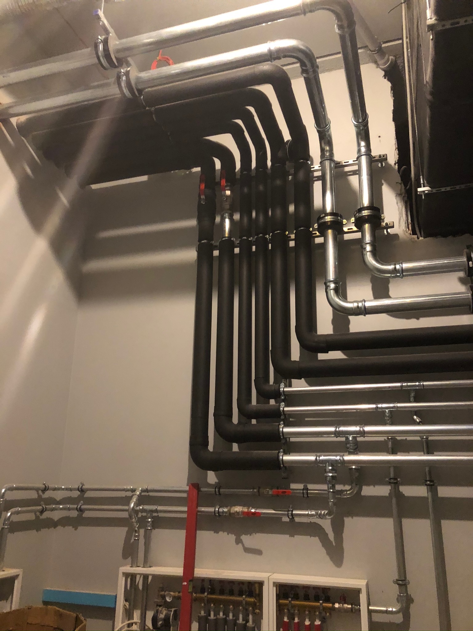 Różne rury: srebrne i czarne, tworzące skomplikowany system instalacji w pomieszczeniu, widoczne zawory i elementy mocujące.