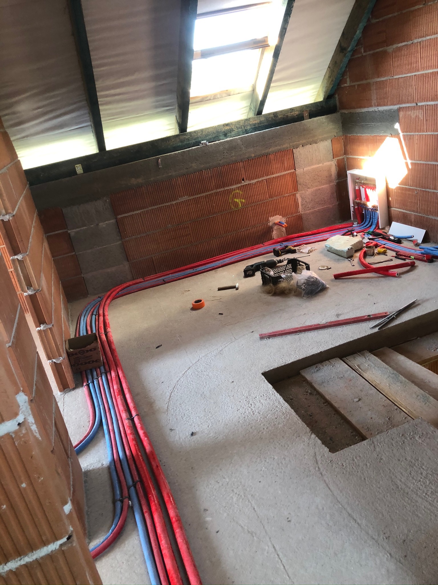 Wnętrze w trakcie budowy z widocznymi, ułożonymi wzdłuż ściany, czerwonymi i niebieskimi rurami instalacji grzewczej i wodnej, narzędziami i skrzynką rozdzielczą na ścianie z cegły.