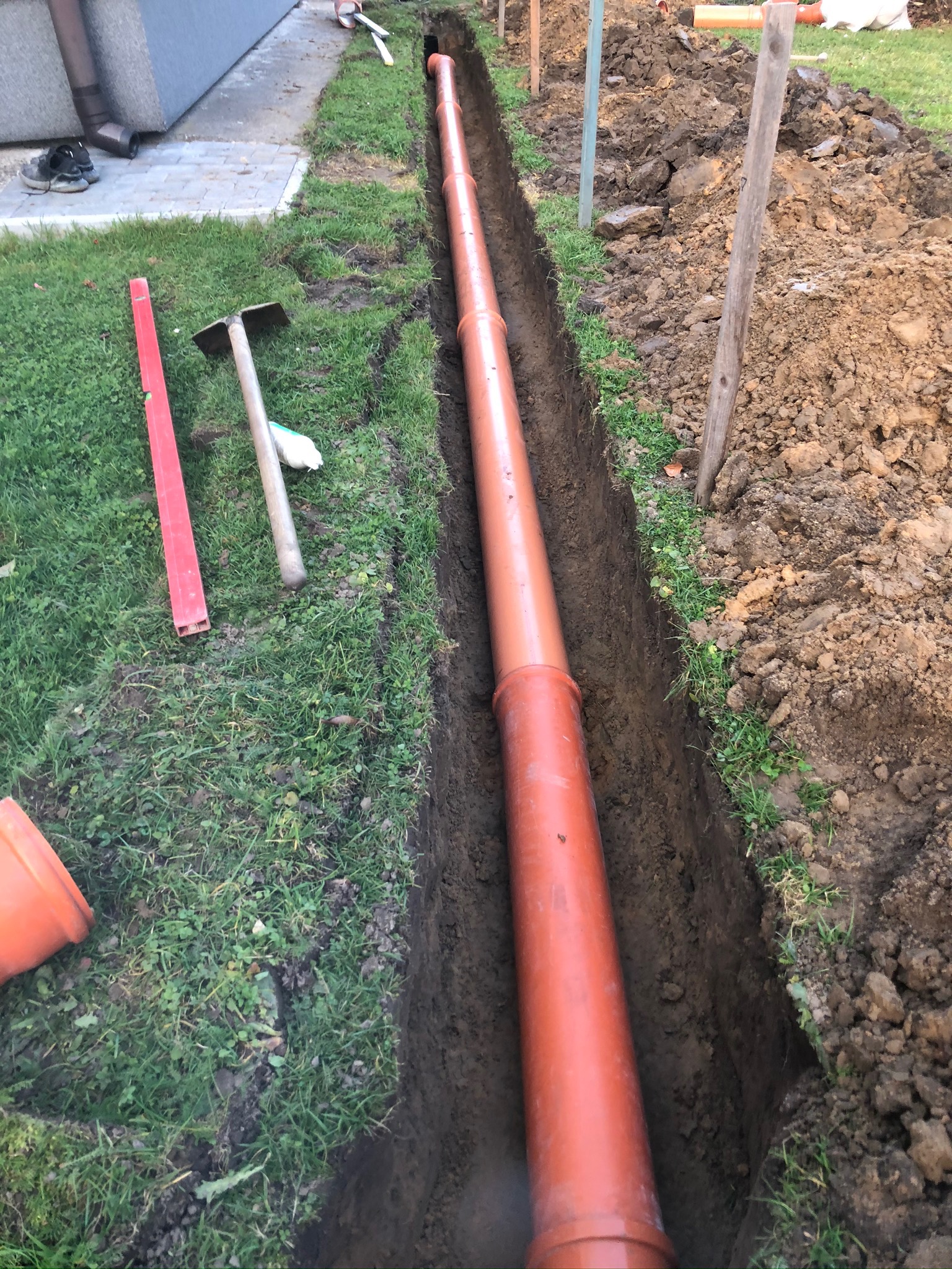Pomarańczowa rura kanalizacyjna ułożona w wykopie ziemnym, widoczny poziomica, szpadel i narzędzia na trawie obok wykopu.