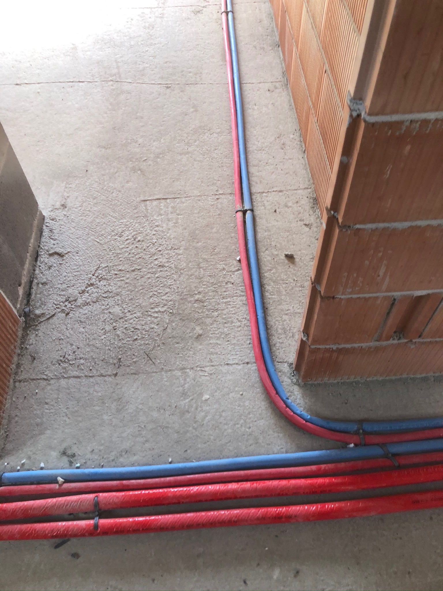 Czerwone i niebieskie rury instalacyjne prowadzone wzdłuż ściany z czerwonej cegły na surowym betonowym podłożu.