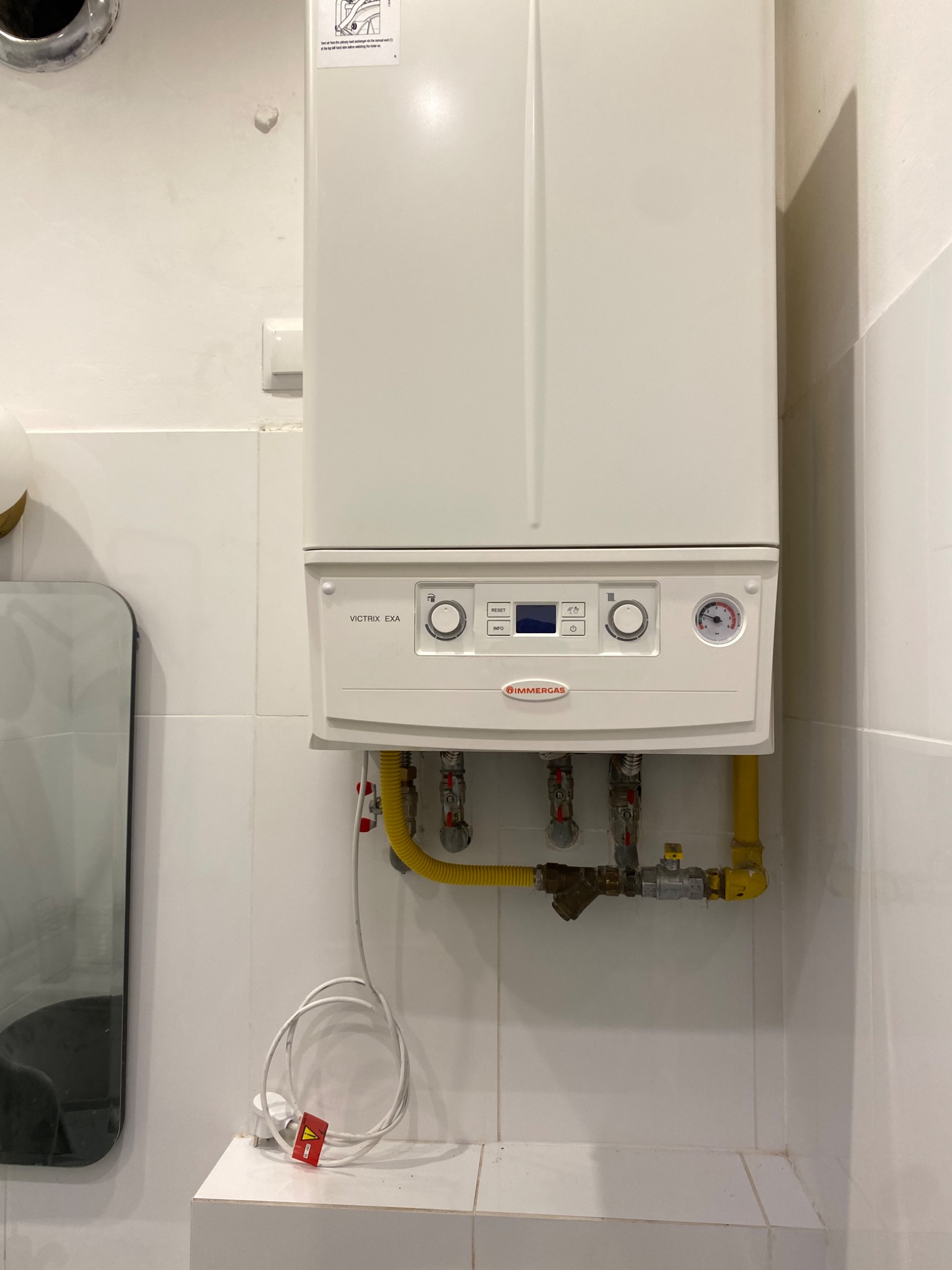 Biały piec gazowy marki Immergas Victrix EXA, zamontowany na białej ścianie z płytek, widoczne żółte rury gazowe i zawory, zasilanie elektryczne z czerwoną etykietą ostrzegawczą, odbicie w lustrze...