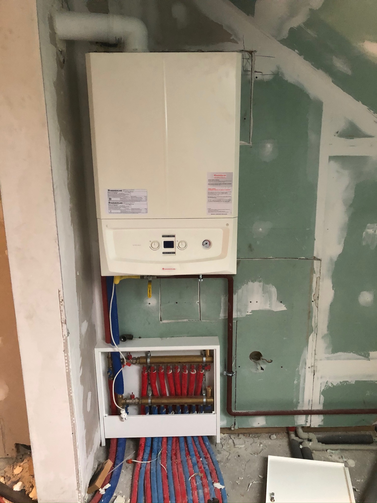 Biały piec gazowy marki Immergas zamontowany na ścianie w surowym stanie, z widoczną instalacją hydrauliczną i rozdzielaczem w białej obudowie, rury czerwone i niebieskie prowadzone po podłodze...