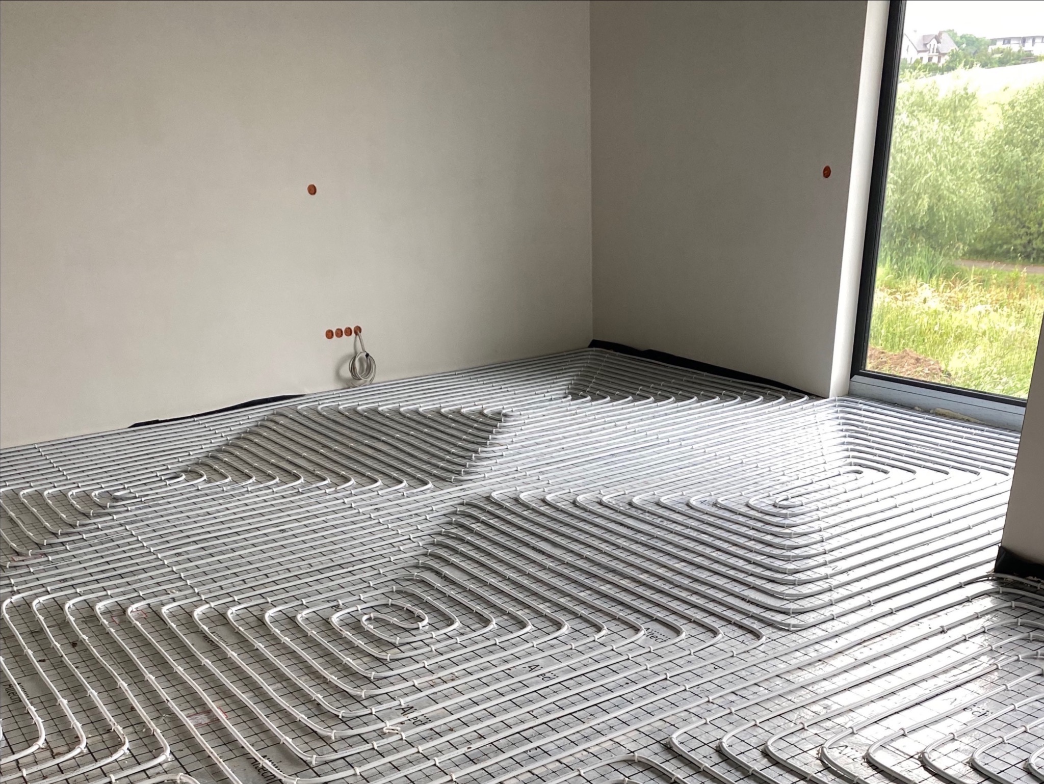 Białe rurki ogrzewania podłogowego ułożone w pętle na szarej macie w surowym pomieszczeniu z dużym oknem wychodzącym na zielony teren.
