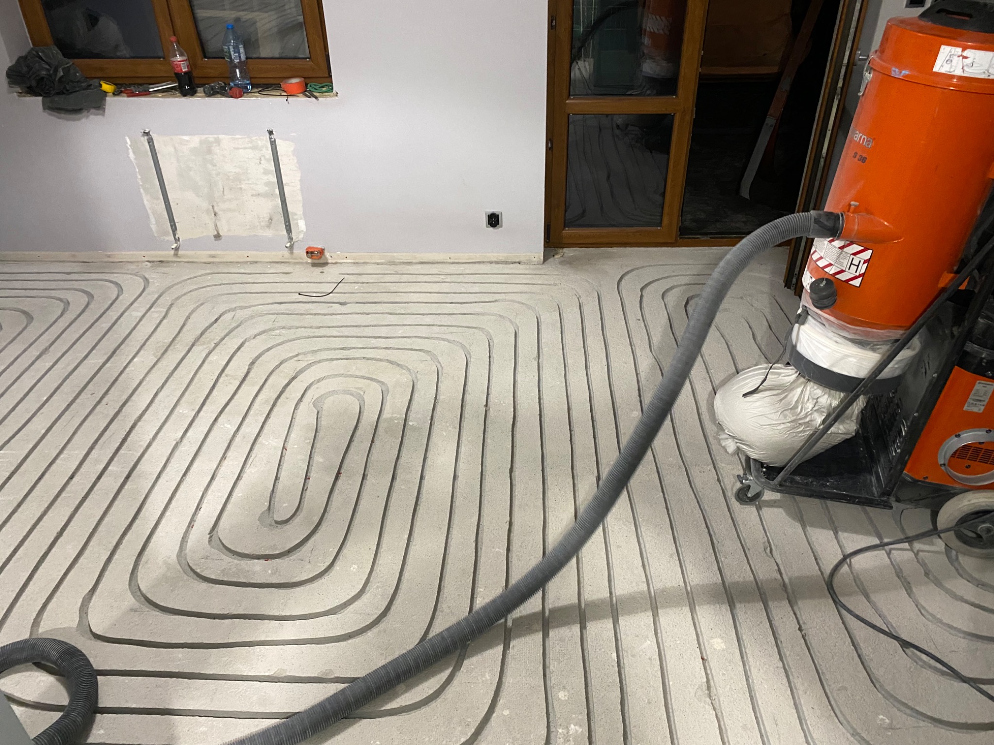 Szare rury ogrzewania podłogowego ułożone w betonie, widoczny pomarańczowy odkurzacz przemysłowy Husqvarna i przygotowane miejsce na grzejnik na ścianie.