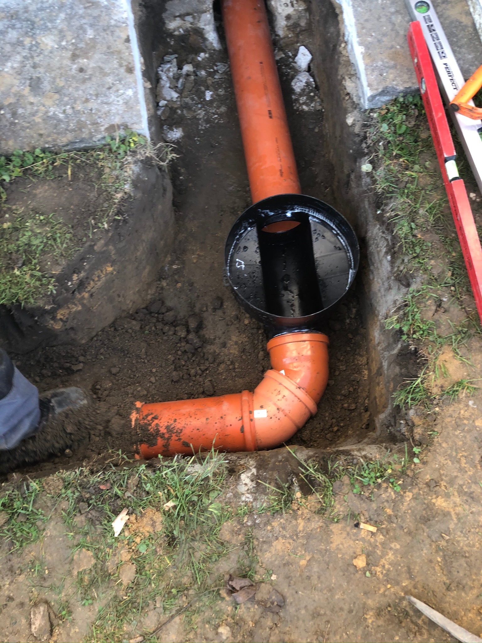 Pomarańczowe rury kanalizacyjne z czarną studzienką rewizyjną w wykopie, widoczny fragment poziomicy i but pracownika.
