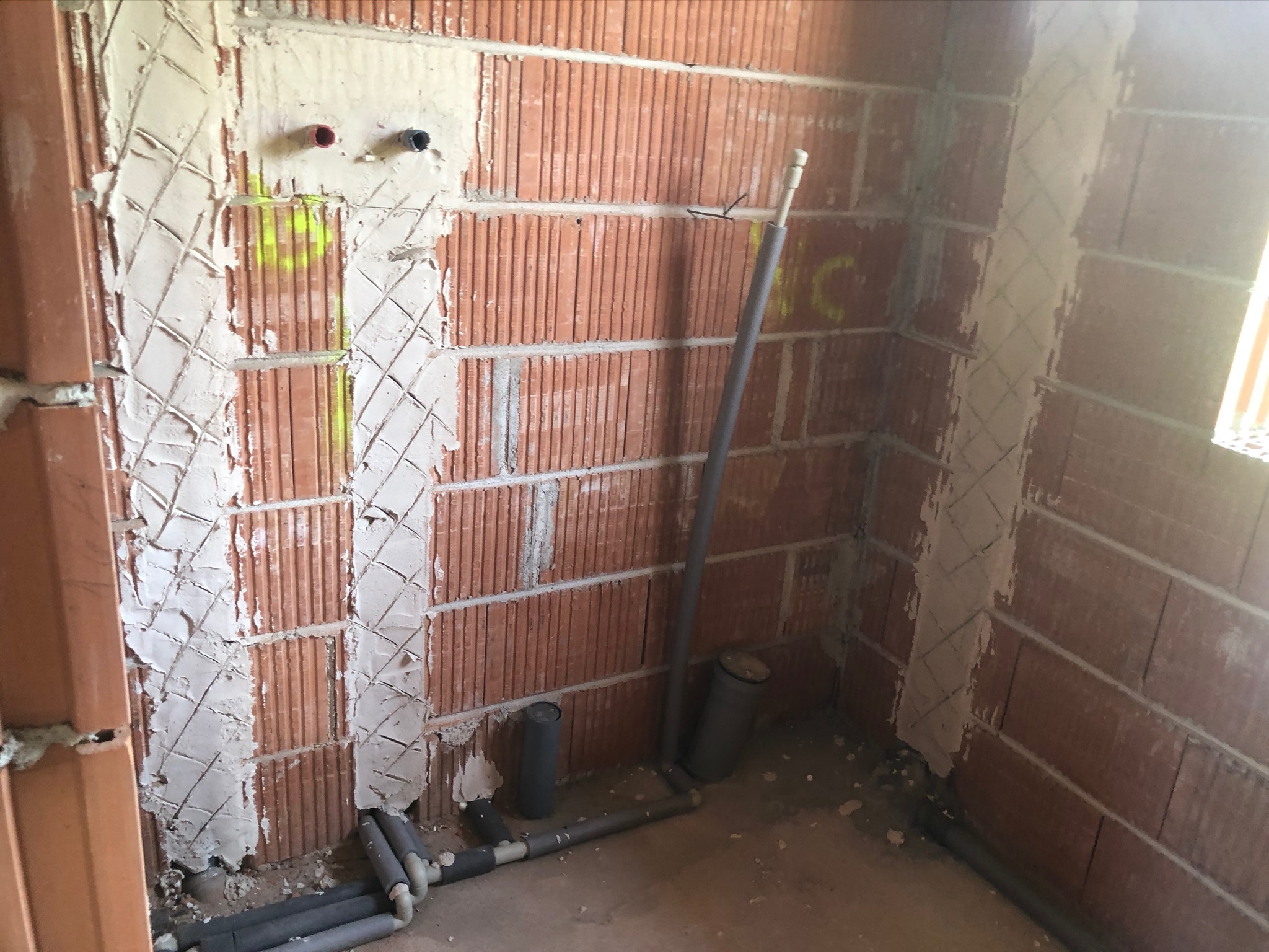 Surowa ściana z czerwonej cegły z wyżłobieniami wypełnionymi zaprawą, widoczne rury instalacji w szarej izolacji i pionowa rura kanalizacyjna, żółte oznaczenia na ścianie.