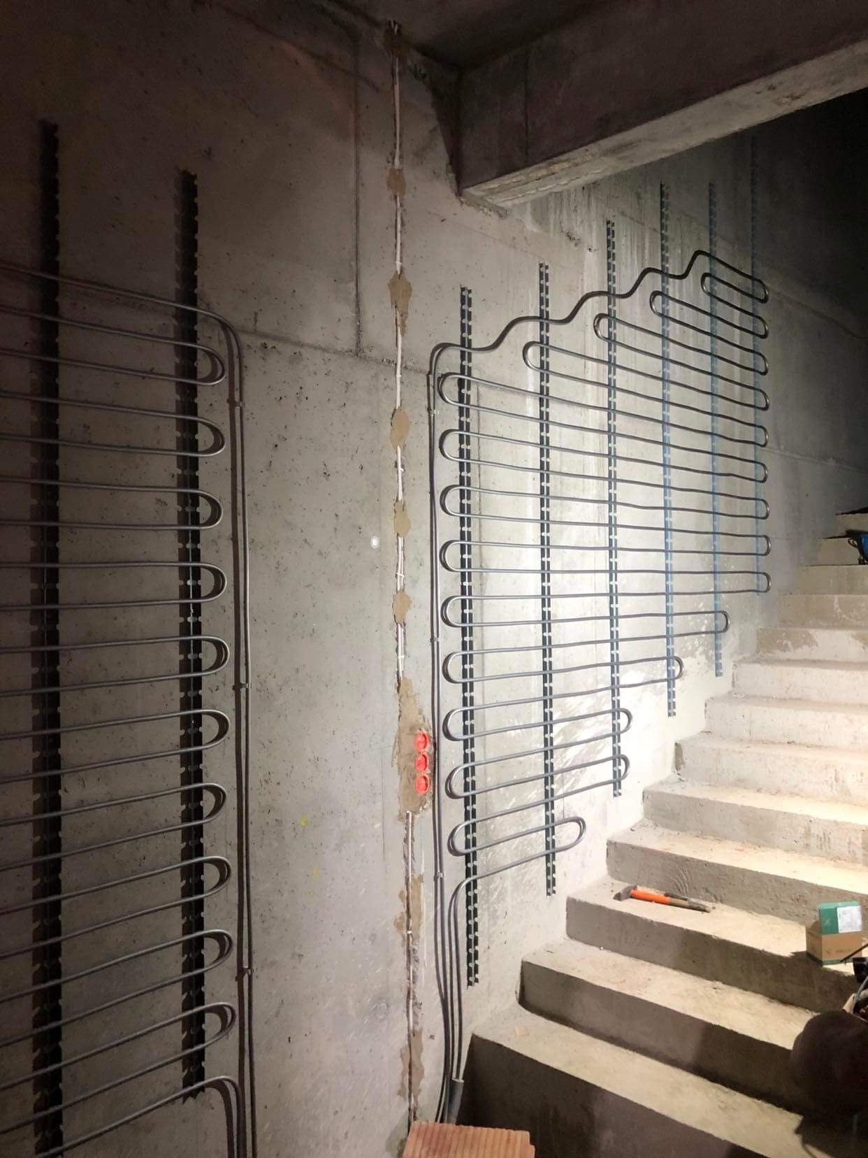 Montaż ogrzewania podłogowego ściennego na surowej betonowej ścianie w klatce schodowej, widoczne mocowania i przewody elektryczne w ścianie, narzędzia leżące na schodach.