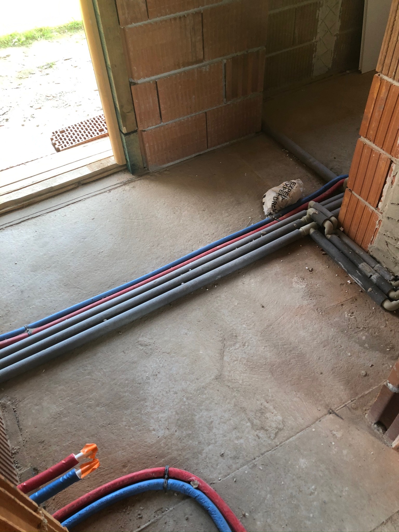 Surowy stan: Rury instalacji wodnej i grzewczej ułożone na betonowej posadzce w nowym budynku z cegły, widoczne izolowane odcinki i końcówki zabezpieczone pomarańczowymi zaślepkami.