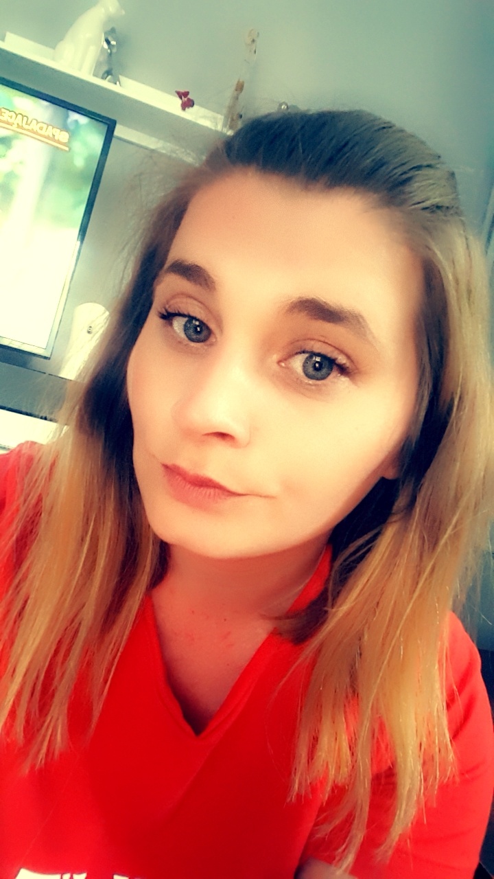 Portret kobiety z blond pasemkami, ubranej w czerwoną koszulkę, z telewizorem i dekoracjami w tle.