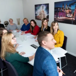 Grupa os&oacute;b uczestniczy w spotkaniu w biurze, na stole leżą dokumenty i szklanki z wodą, w tle wiszą obrazy z architekturą miejską.