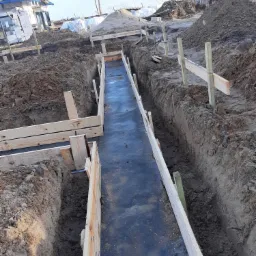 Wykonanie wykopu pod fundamenty z szalunkiem drewnianym i folią hydroizolacyjną, widoczne pryzmy ziemi i budynki w tle.