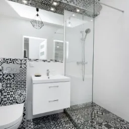 Nowoczesna łazienka z kabiną prysznicową typu walk-in, białą szafką pod umywalką, wiszącą toaletą i czarno-białymi płytkami w stylu patchwork na podłodze i ścianie za toaletą, z geometryczną lampą...
