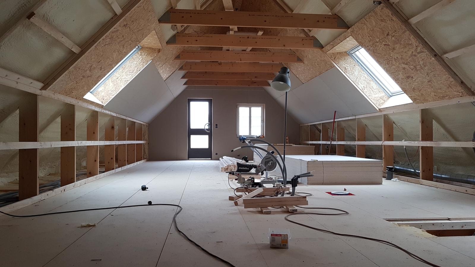 Adaptacja poddasza: widok na drewnianą konstrukcję dachu z oknami połaciowymi, ocieplone ściany pianką poliuretanową, szkielet regału, narzędzia do cięcia drewna i stos płyt kartonowo-gipsowych.