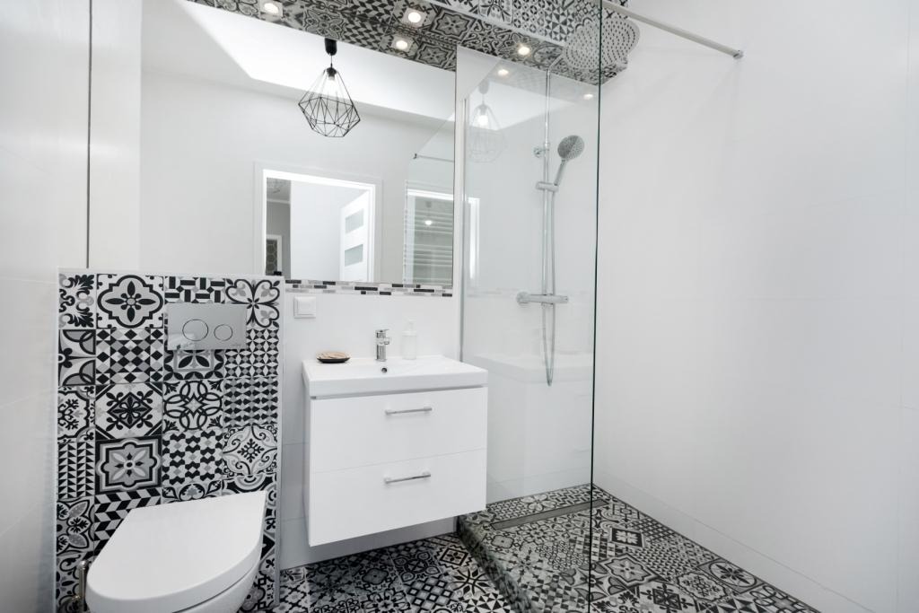 Nowoczesna łazienka z kabiną prysznicową typu walk-in, białą szafką pod umywalką, wiszącą toaletą i czarno-białymi płytkami w stylu patchwork na podłodze i ścianie za toaletą, z geometryczną lampą...