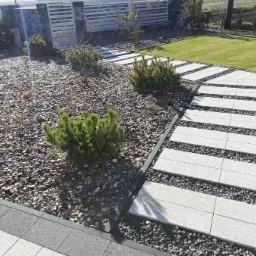 Ścieżka z jasnych, prostokątnych płyt ułożona w trawniku, otoczona szarym żwirem i niskimi roślinami iglastymi, widok na zadbany ogród.