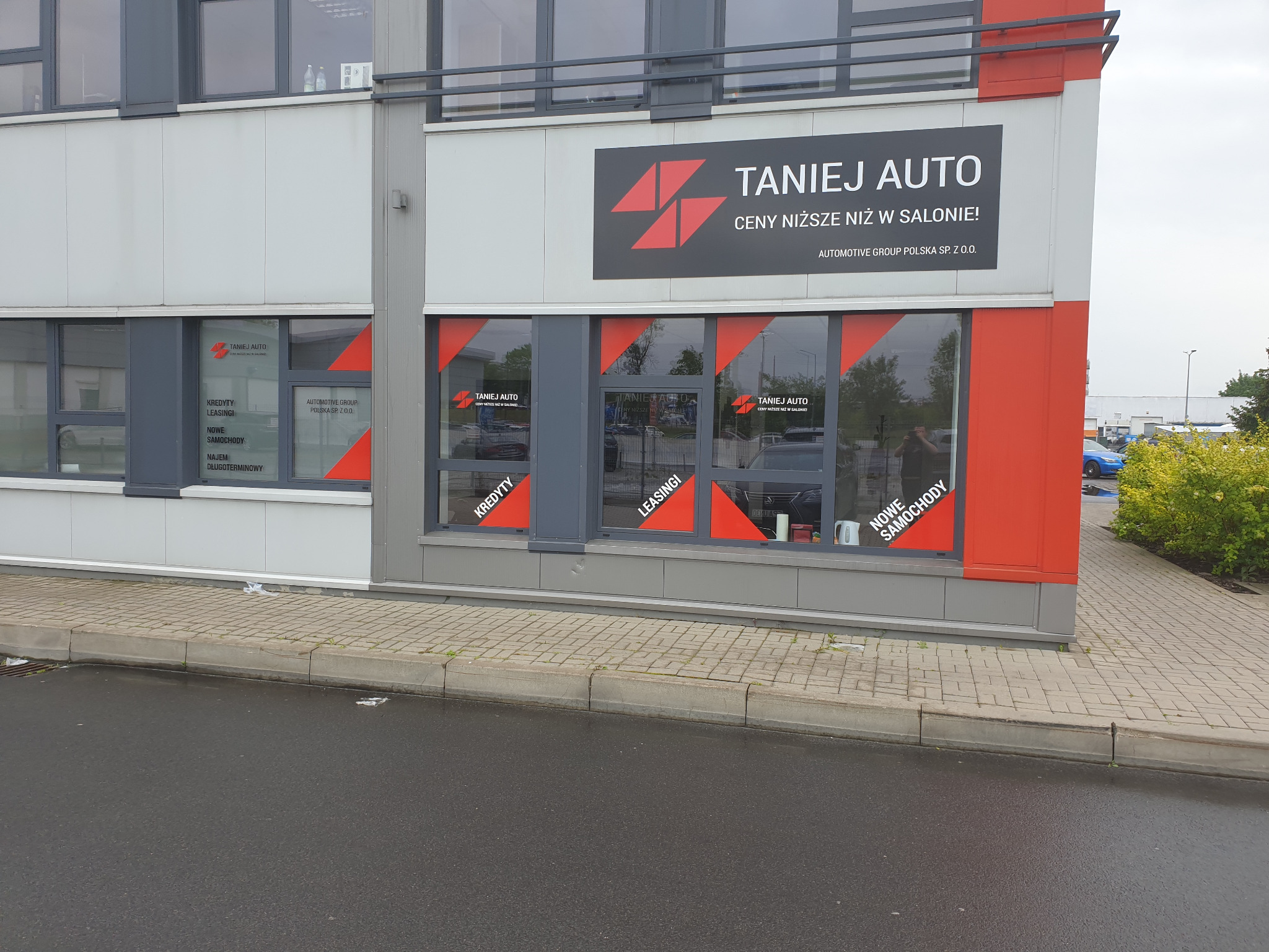 Budynek firmy z szyldem 'TANIEJ AUTO', okna z naklejkami informującymi o kredytach, leasingu i nowych samochodach, widok z zewnątrz z chodnika i ulicy.