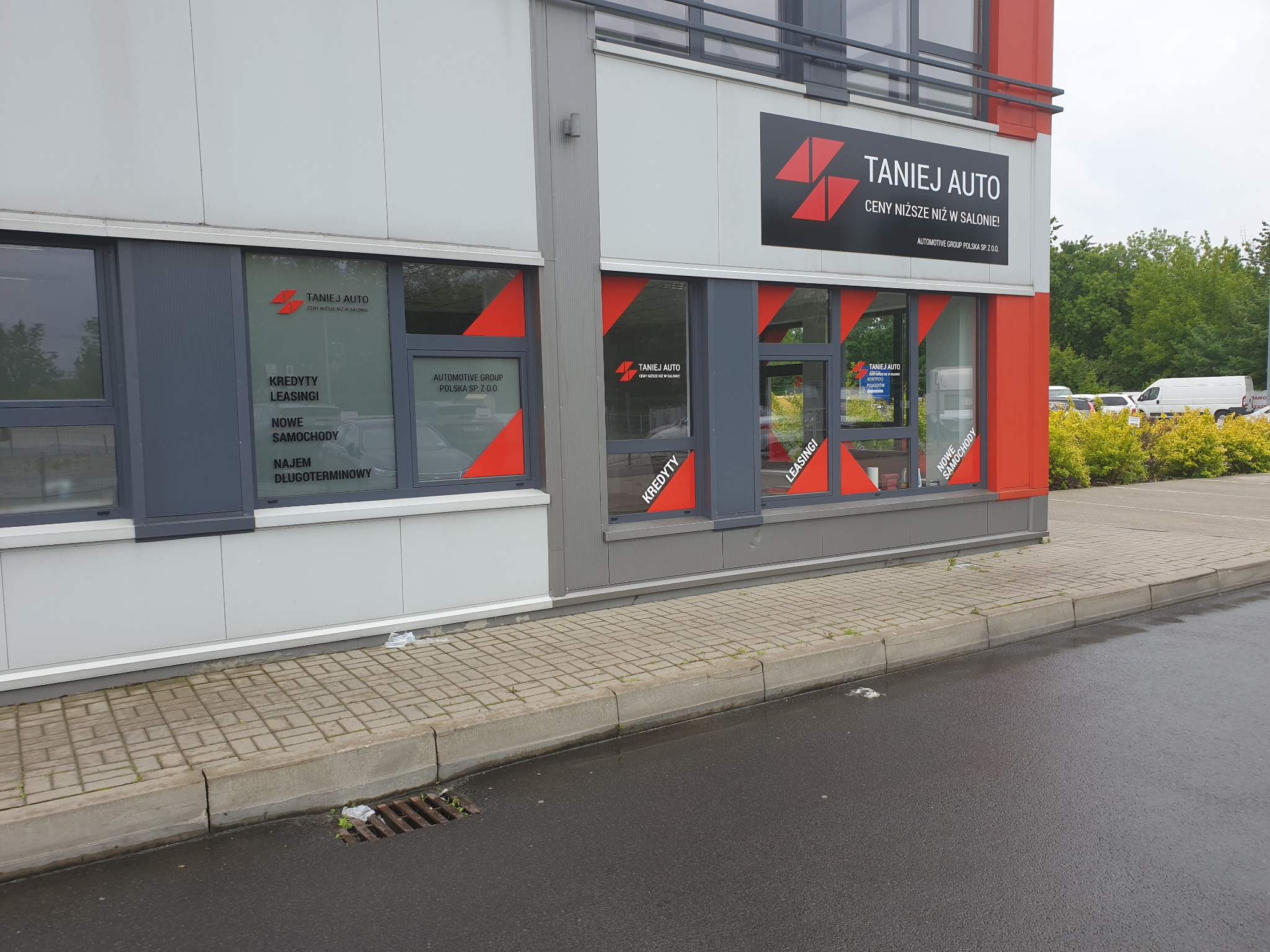 Fasada budynku firmy 'Taniej Auto' z widocznymi oknami, na których umieszczone są informacje o kredytach, leasingu, nowych samochodach i najmie długoterminowym, z logo firmy na górze i czerwonymi...