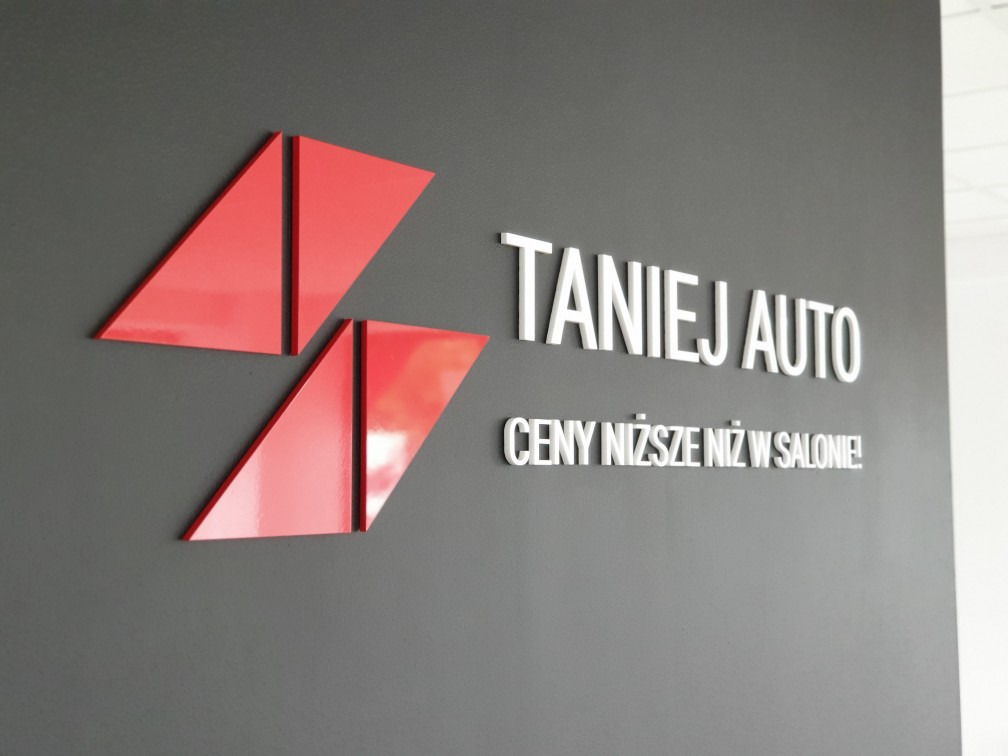 Logo firmy 'Taniej Auto' z czerwonym, geometrycznym symbolem i hasłem 'Ceny niższe niż w salonie!' na szarej ścianie.