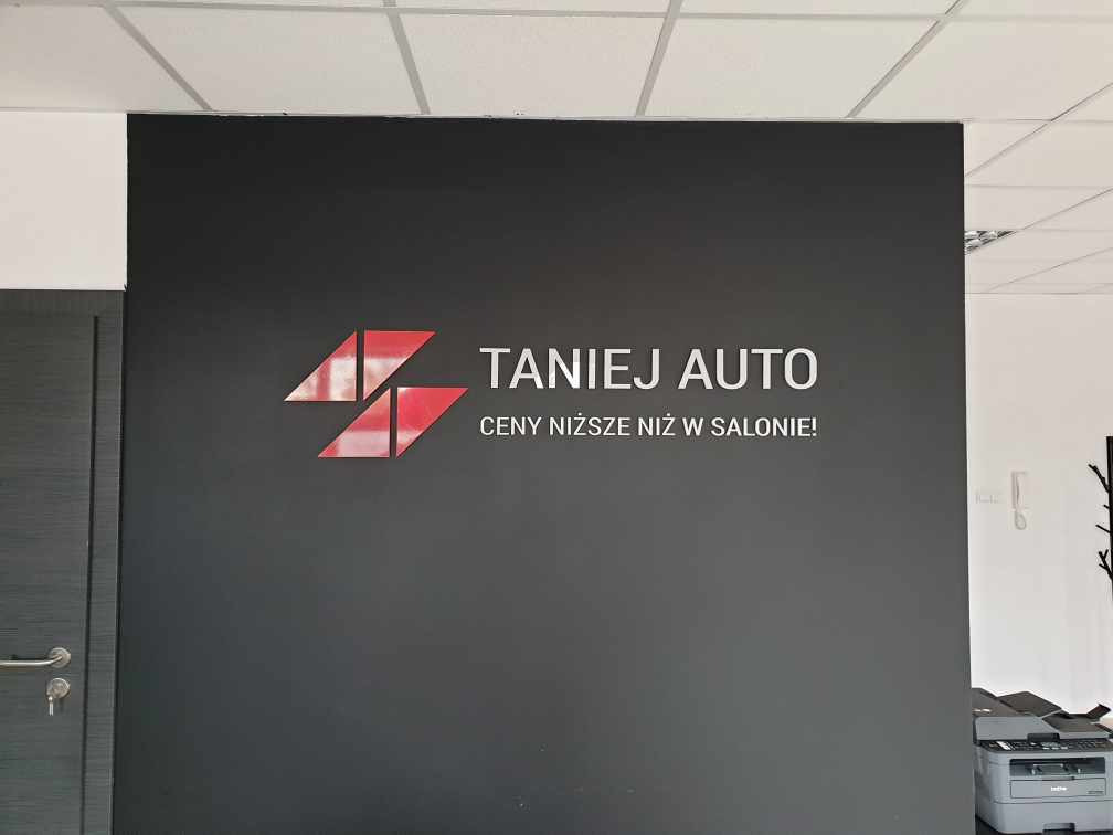 Logo firmy 'Taniej Auto' na ciemnoszarej ścianie z hasłem 'Ceny niższe niż w salonie!', widoczny fragment drzwi po lewej i drukarka po prawej.